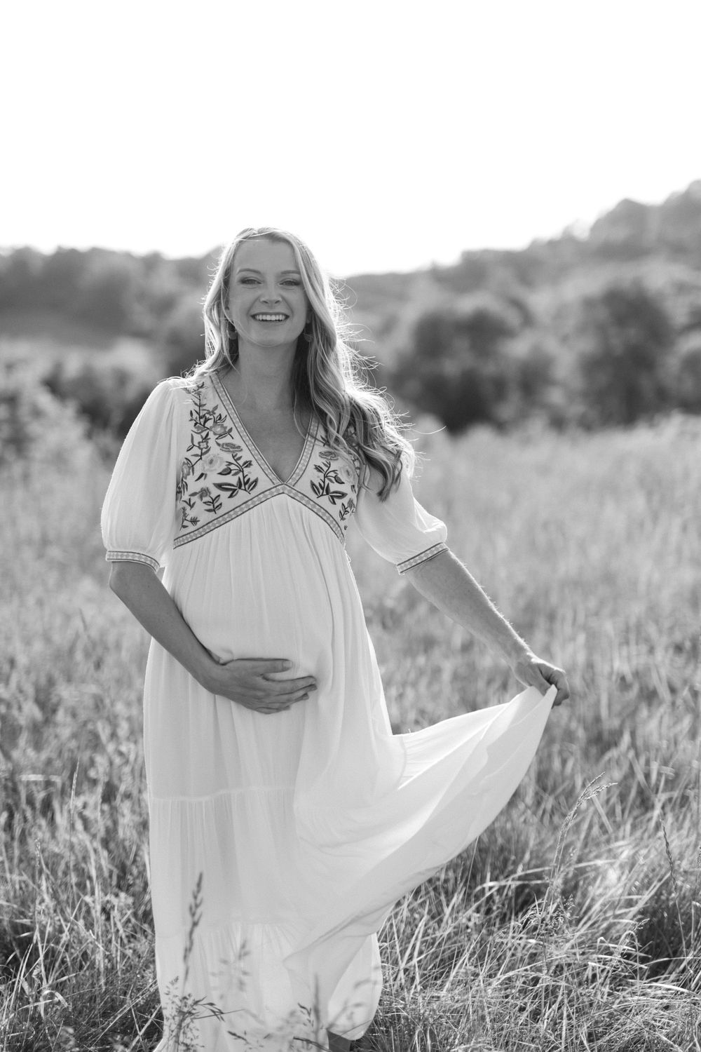 Smiling pregnant woman in white embroidered boho dress holding belly in sunny field.