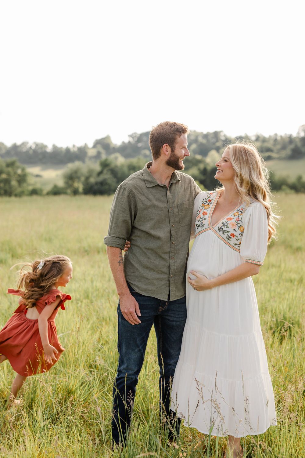 Pregnant couple sharing a smile in green field while young daughter in red dress twirls nearby.