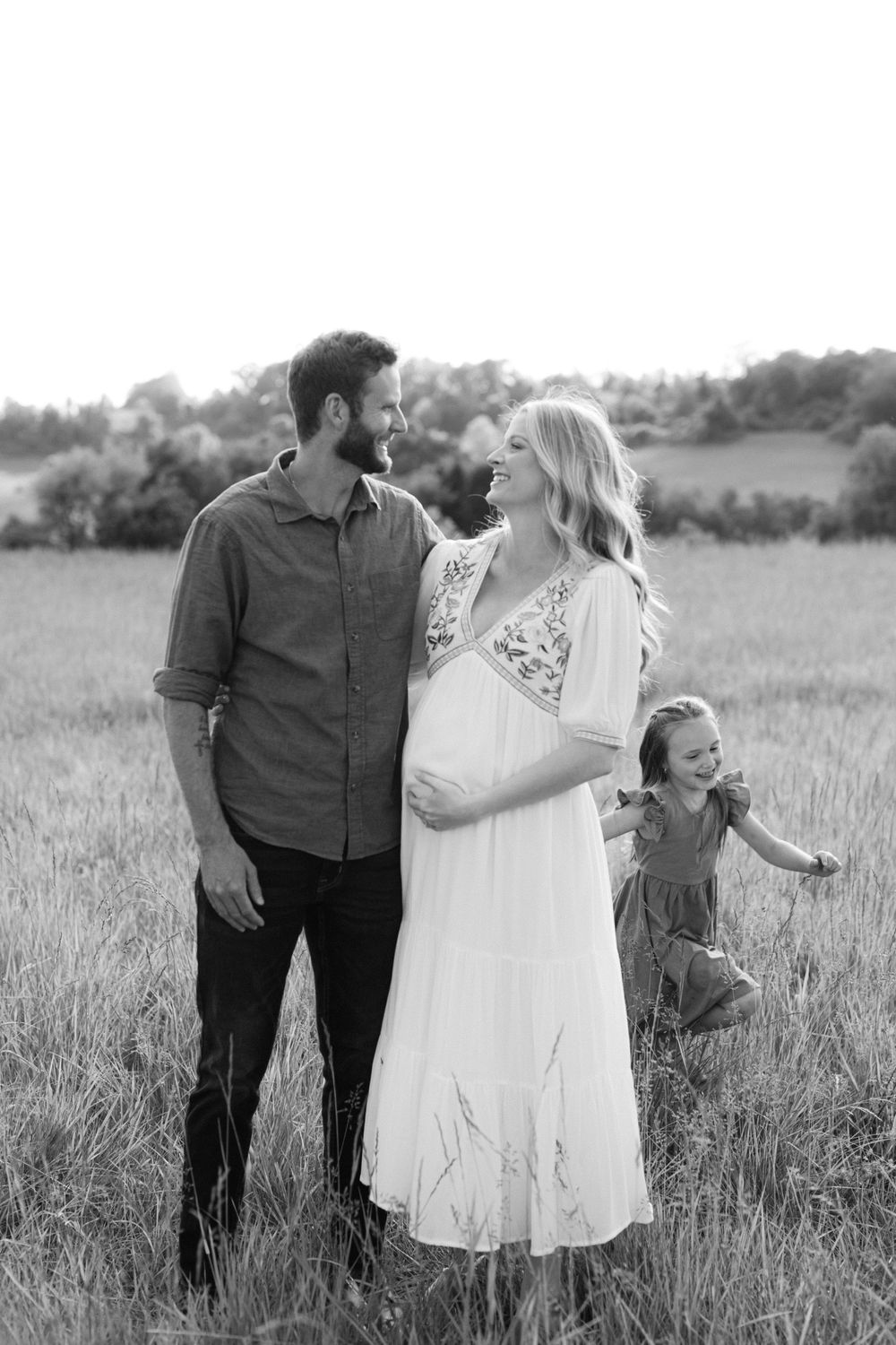 Black and white maternity photo of couple gazing at each other as daughter plays behind them.