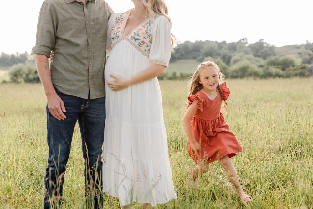 Pregnant woman in white embroidered dress stands with husband as daughter dances beside them.