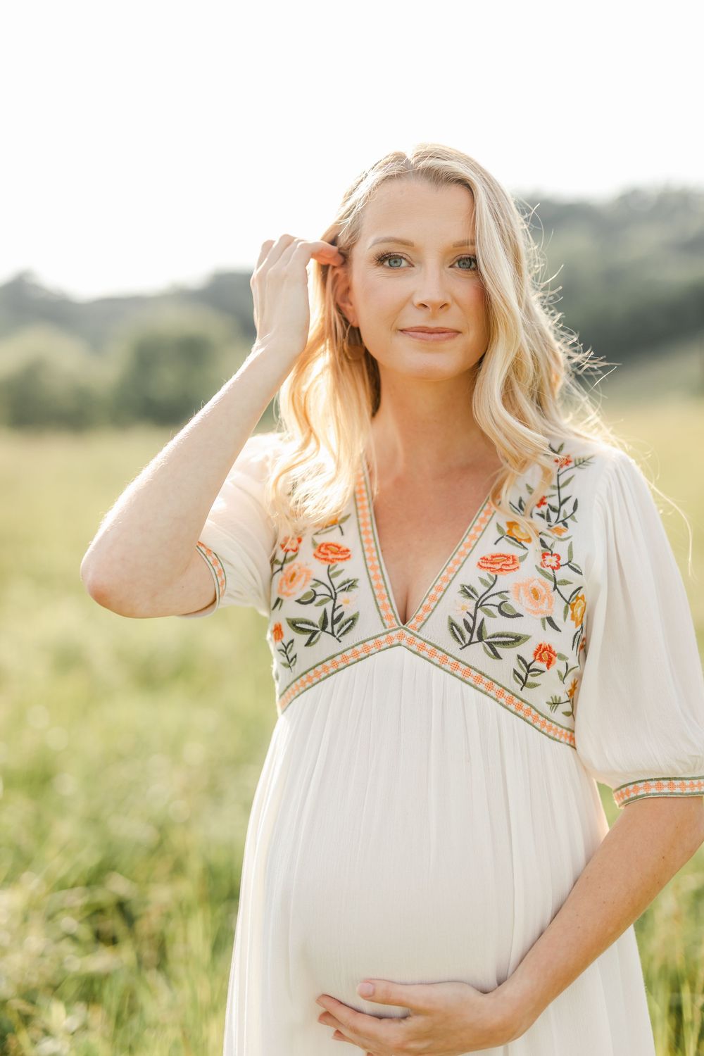 Pregnant blonde woman in white floral dress gently touching hair in a sunlit green field.