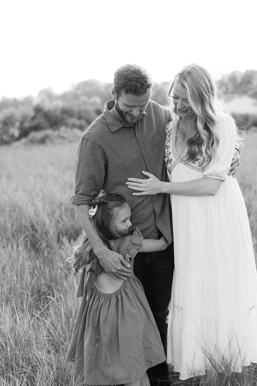 Black and white photo of parents and young daughter embracing in a meadow.