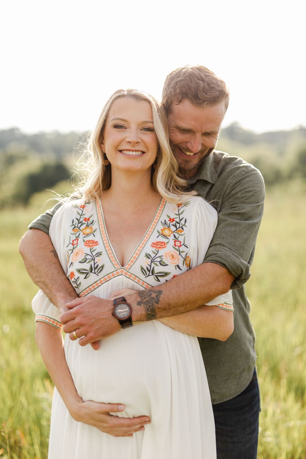 Smiling pregnant woman in floral white dress embraced by bearded man in green shirt in a field.