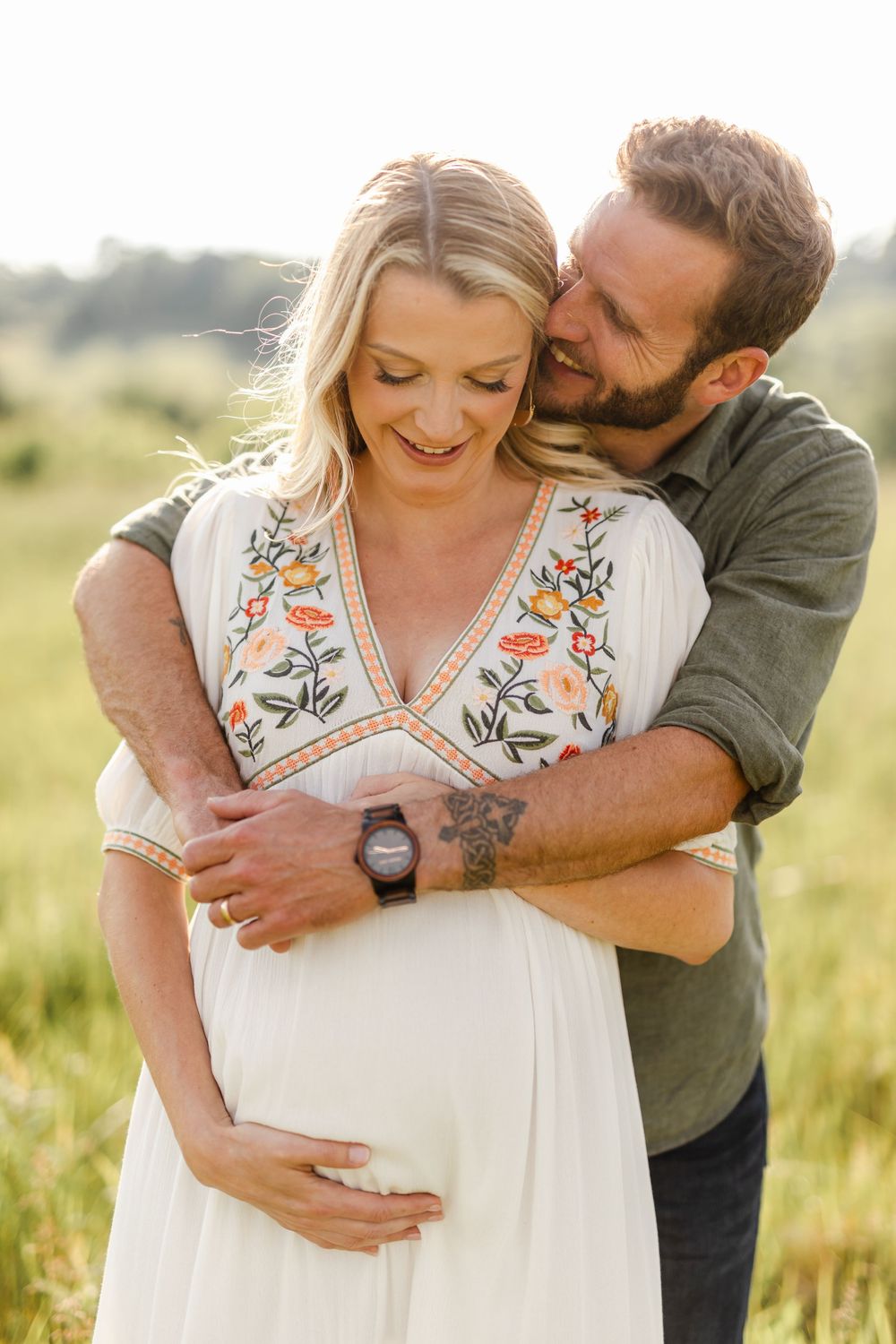 Couple laughing together outdoors, man with tattoo embracing pregnant woman in embroidered white dress.