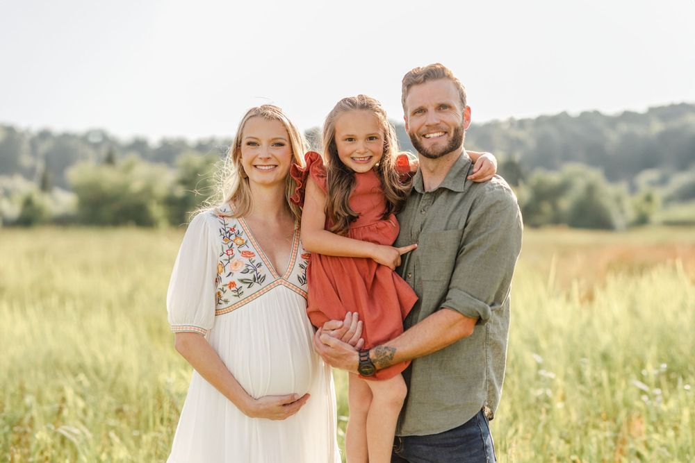 Happy family of three in sunny field, pregnant mom, smiling dad holding young daughter in red dress.