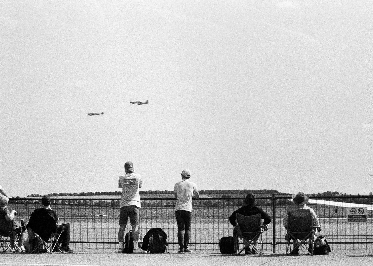 People watching military aircraft fly overhead at an outdoor airshow, standing and sitting in folding chairs near a fence.