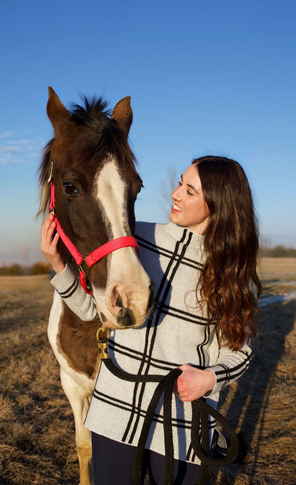 Young woman with long brown hair smiling while petting a brown and white horse wearing a red halter outdoors.