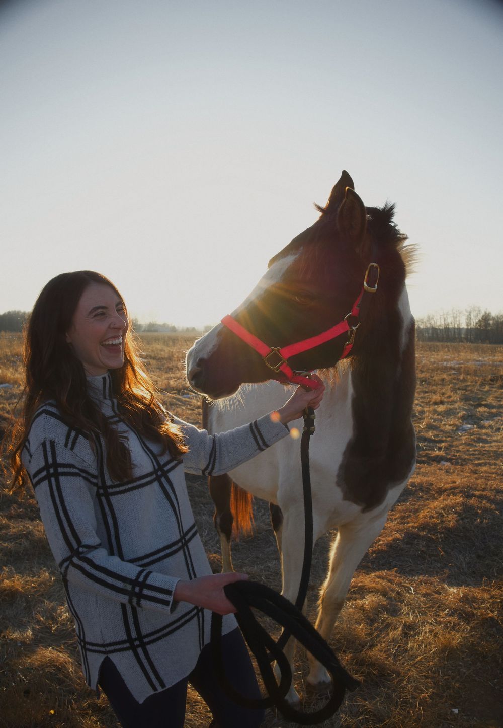 A smiling young woman with long brown hair holds the reins of a brown and white horse wearing a red halter outdoors.
