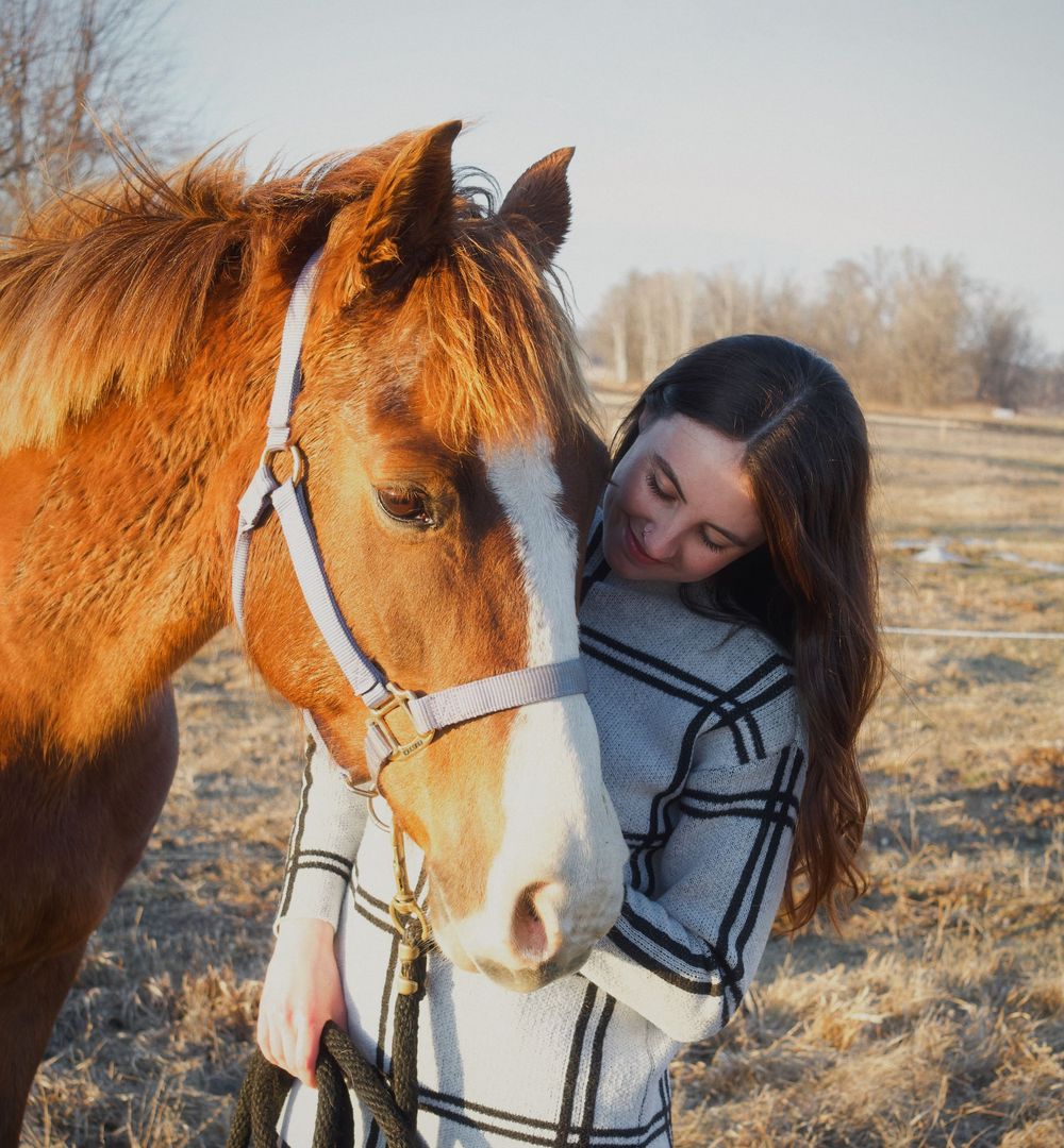 Young woman with long brown hair affectionately embracing a chestnut horse wearing a white halter in a field.