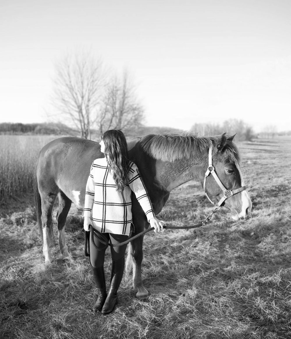 Black and white photo of a woman standing with two horses in a field, gently touching one horse on a calm day.