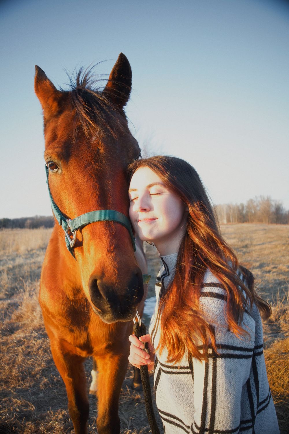 Young woman with red hair smiling peacefully beside a chestnut horse in a golden field at sunset.