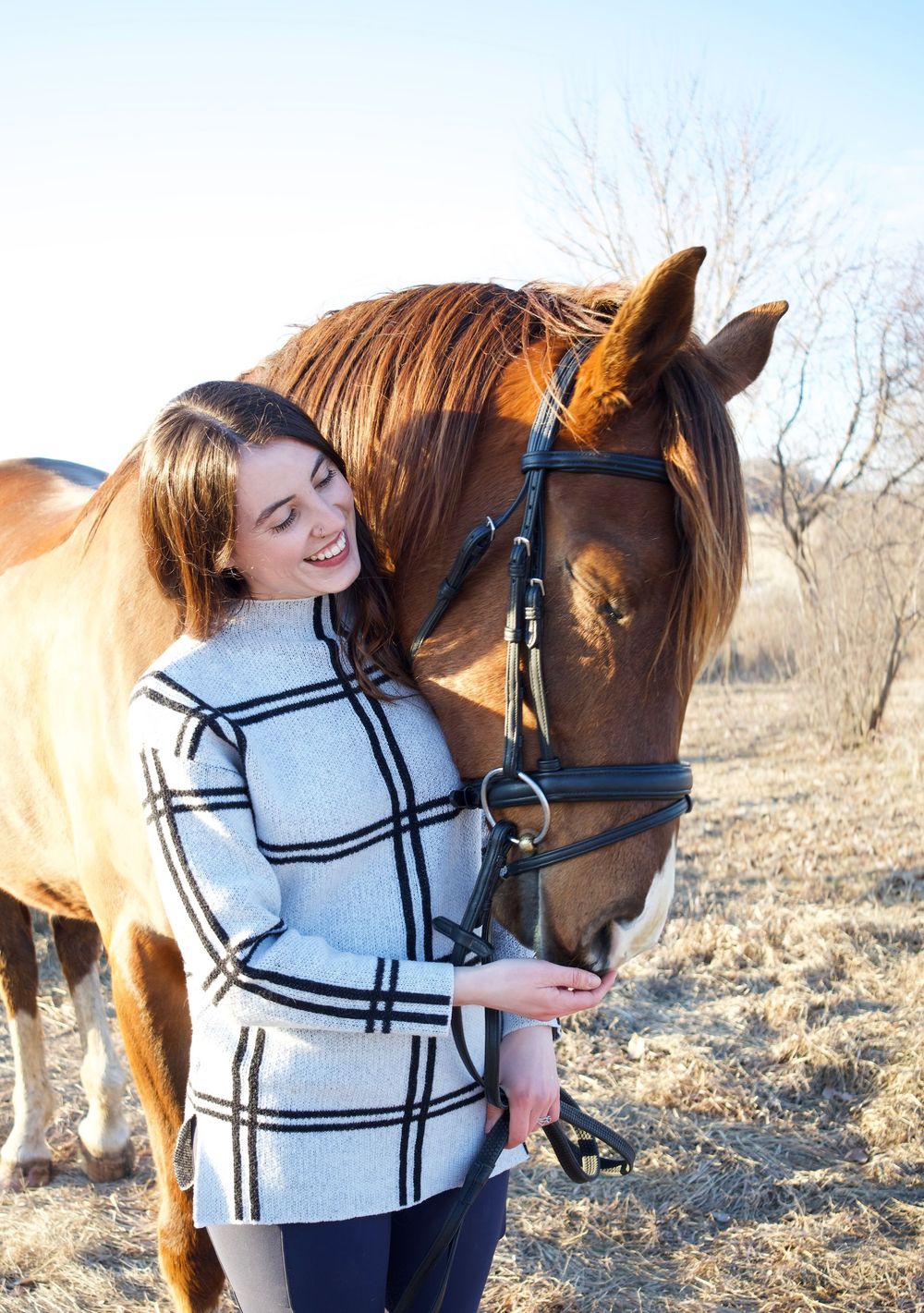 Smiling young woman in plaid coat feeding a chestnut horse with a black bridle outdoors in winter.