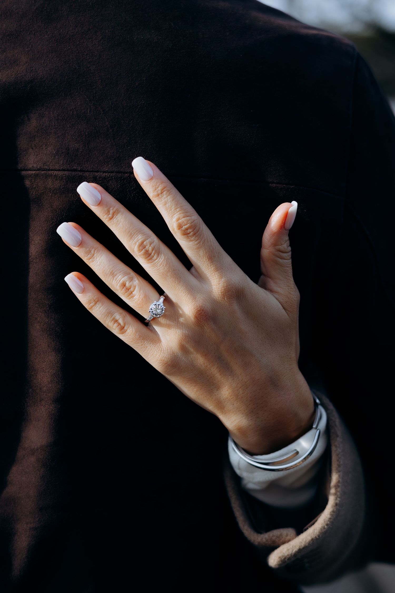 A hand with nude nails displays a sparkling diamond engagement ring against a dark brown jacket in bourton on the water Cotswolds.