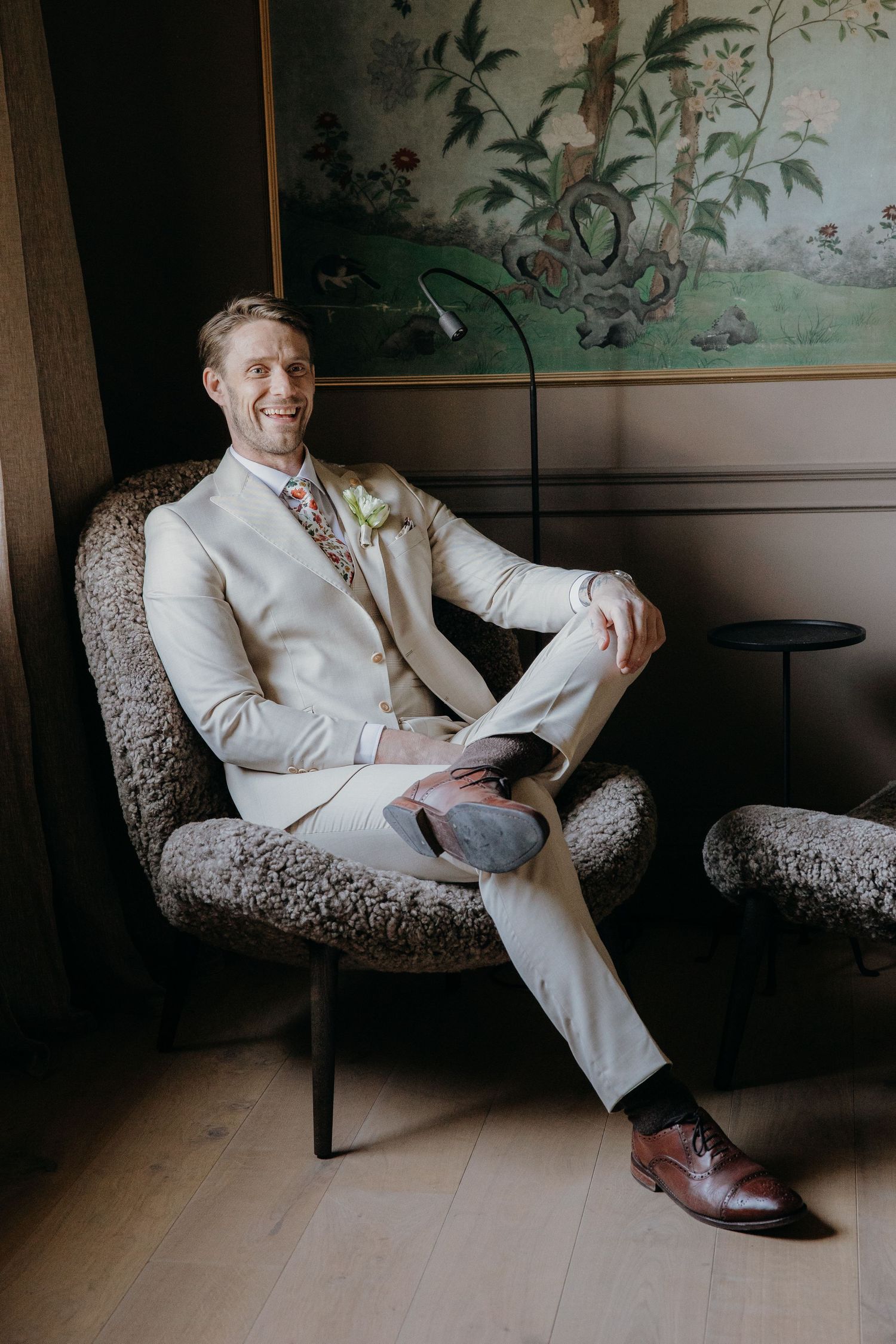 A smiling groom in a cream suit with floral boutonniere sits relaxed in a textured armchair, wearing brown oxford shoes.