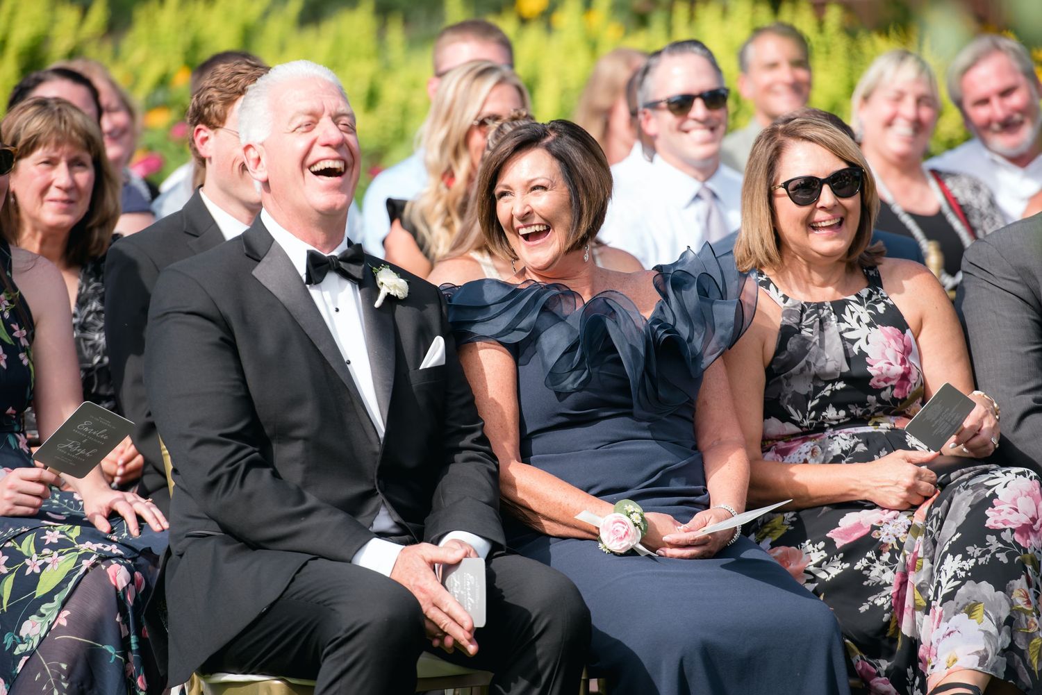 Guests laughing joyfully at an outdoor wedding ceremony, dressed in formal attire including tuxedo and navy gowns.