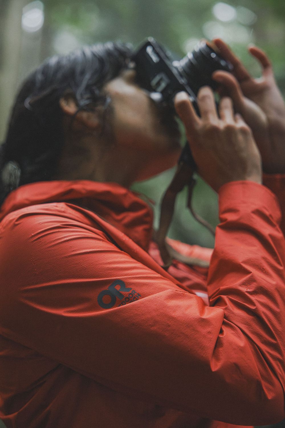 Person in red jacket photographing upward with a film camera in a forest setting.