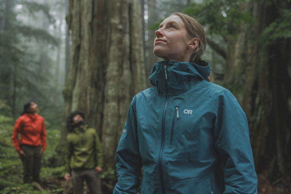Woman in teal Outdoor Research rain jacket gazing up in misty forest with hikers in background.
