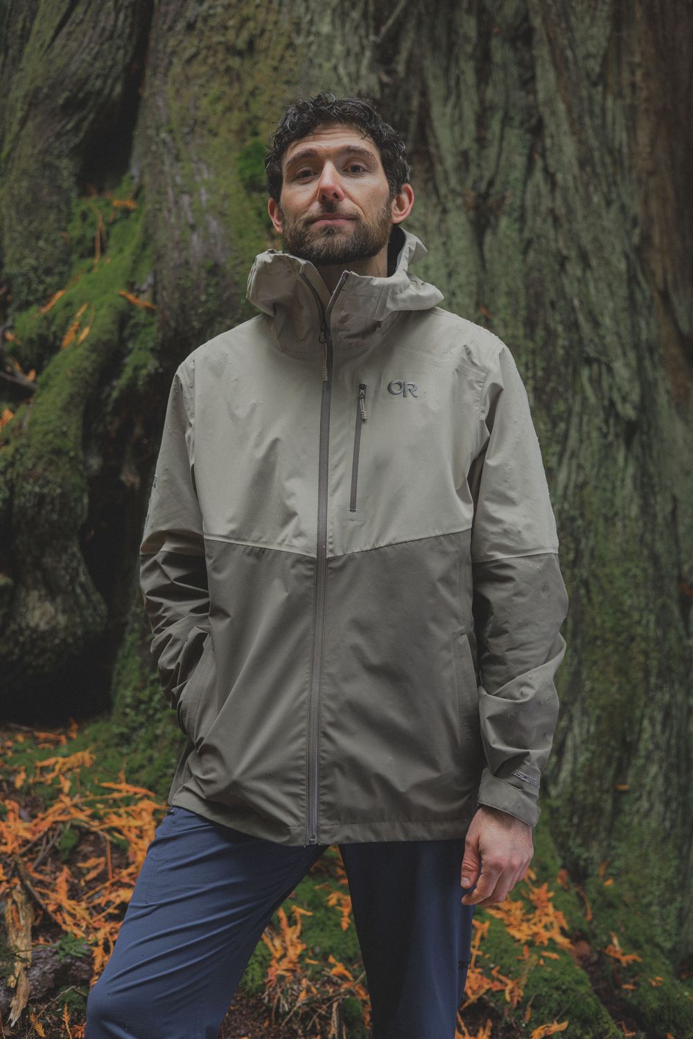 Man wearing a tan hooded outdoor rain jacket standing in a mossy forest setting.