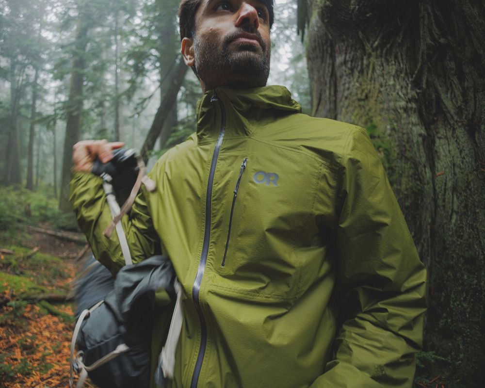 Man wearing olive green rain jacket hiking in misty forest, carrying backpack and trekking pole.