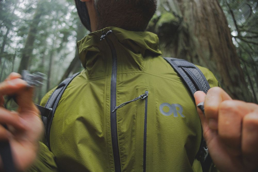 Hiker wearing olive green Outdoor Research rain jacket with backpack in lush forest setting.