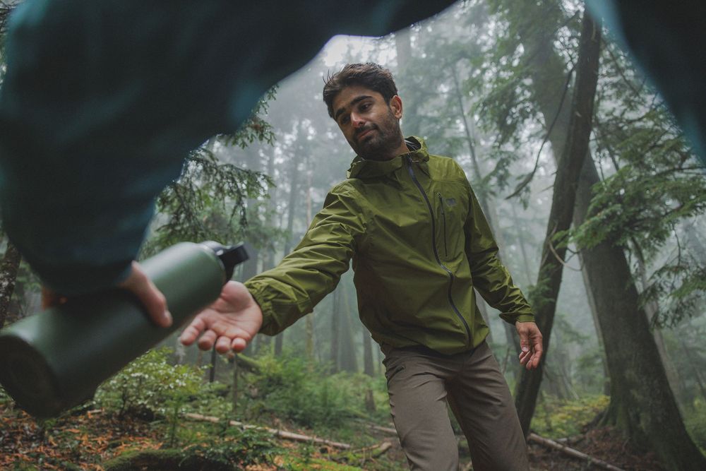 Hiker in green jacket reaching for a water bottle in a misty forest during an outdoor adventure.