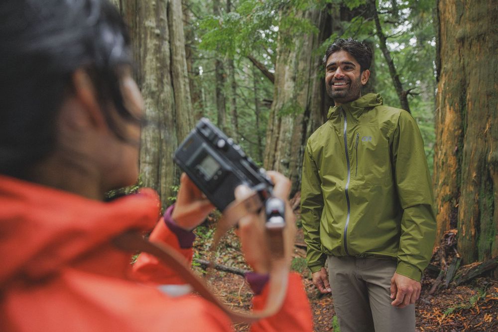 Person in red jacket photographs smiling man in green rain jacket standing among tall trees in a lush forest.