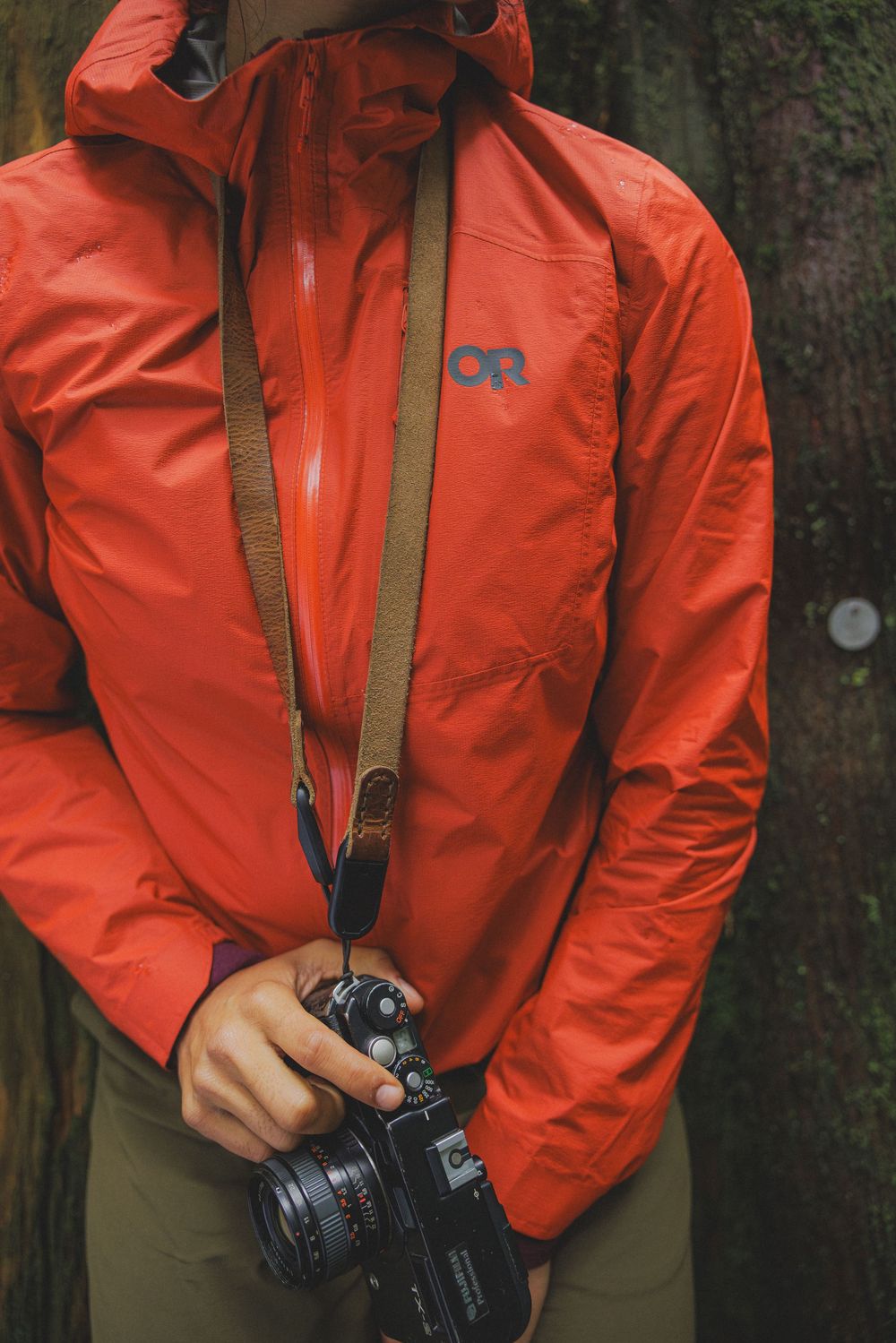 Person wearing an orange Outdoor Research jacket holding a film camera with a brown leather strap outdoors.