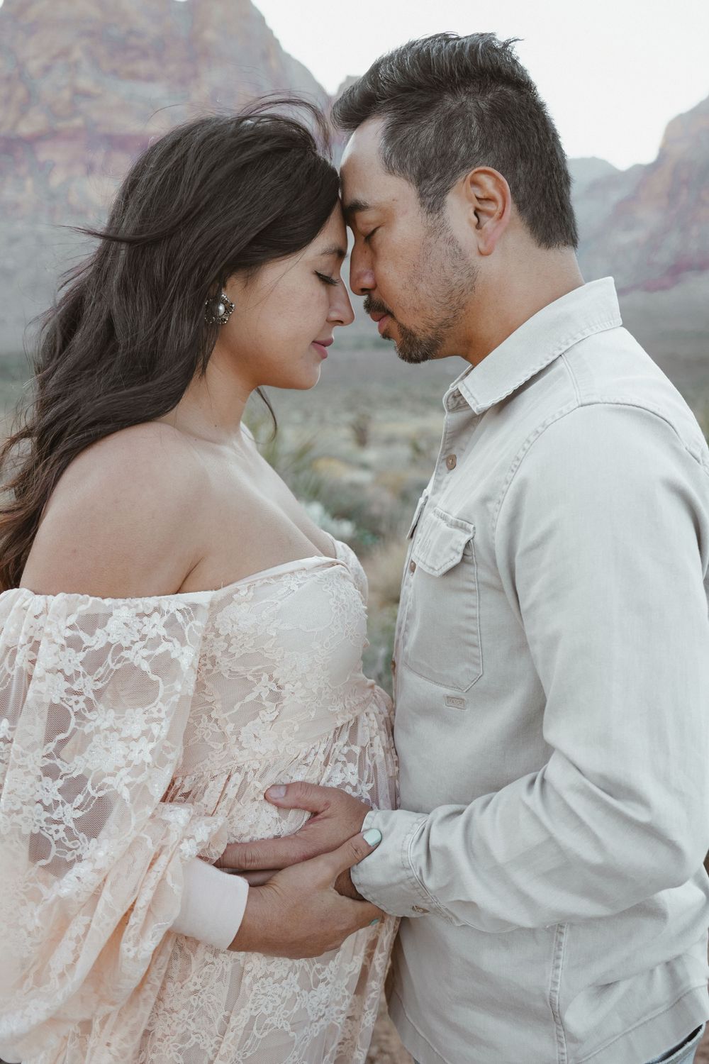 Couple sharing an intimate moment outdoors, pregnant woman in lace dress with partner holding her belly, red rock canyon backdrop.