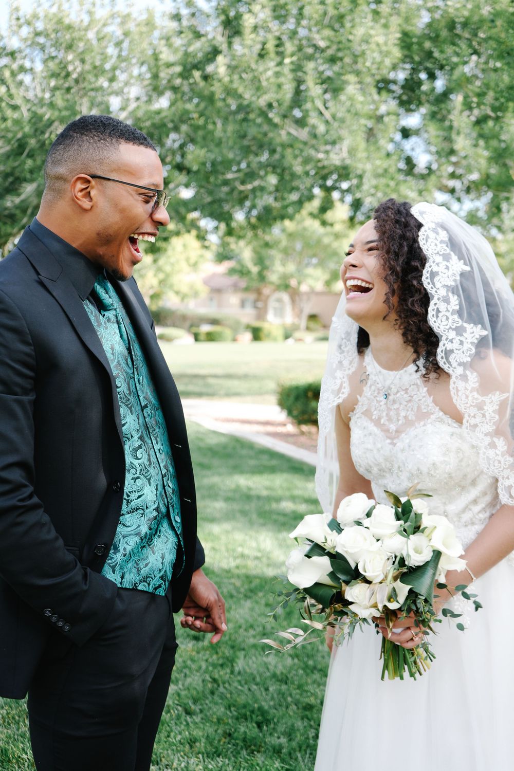 A laughing bride in a white lace gown and veil holds a bouquet while a groom in a black suit with teal vest smiles.