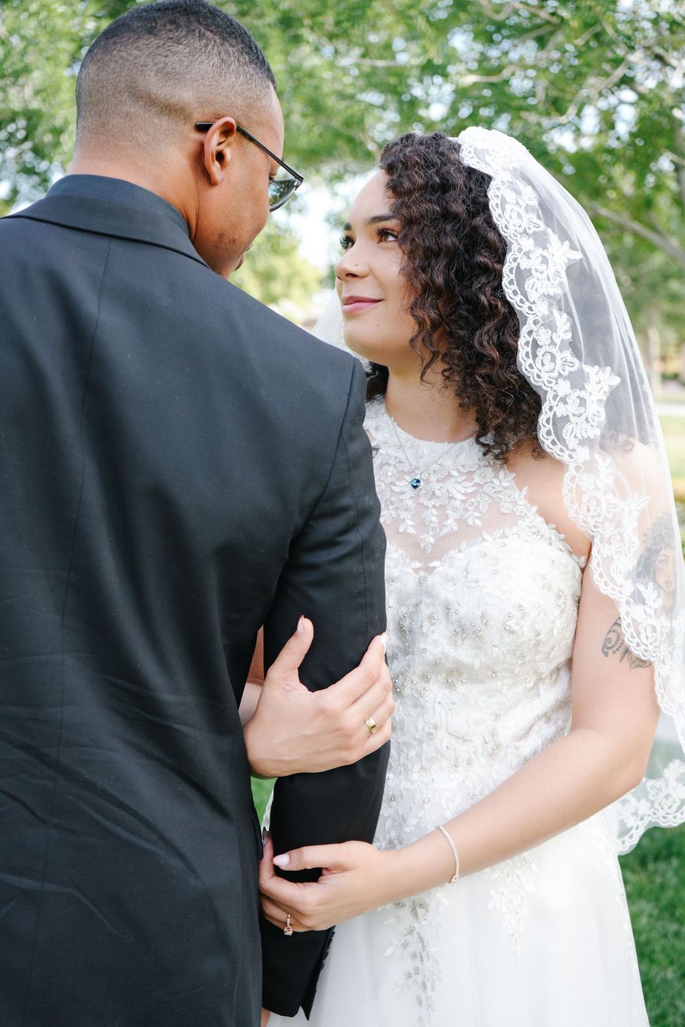 A bride in a lace wedding dress and veil gazes lovingly at her groom in a black suit outdoors.