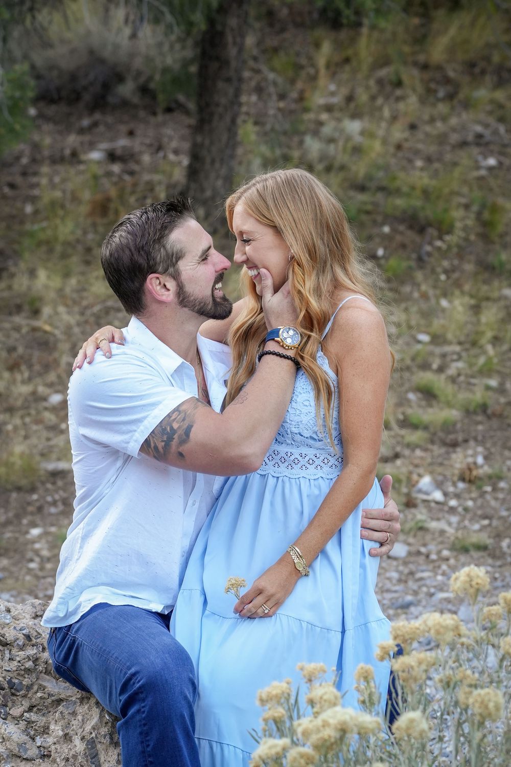 A couple shares a romantic moment outdoors, the woman in a blue dress and man in white shirt embracing near wildflowers.