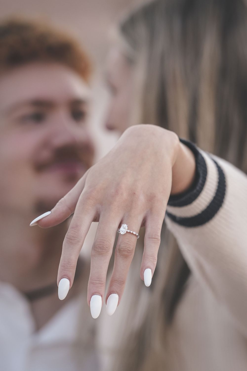 Woman showing off diamond engagement ring with white almond nails, smiling man with red hair in background.