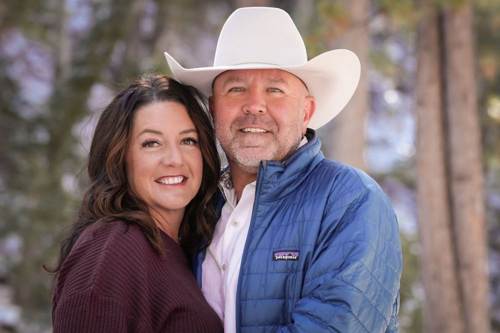 A smiling couple poses outdoors, man wearing a white cowboy hat and blue jacket, woman in burgundy top.