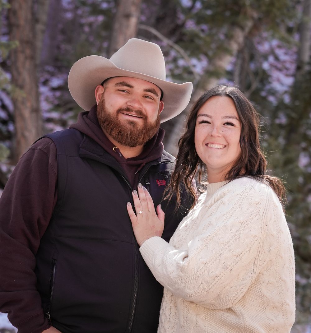 A smiling couple poses outdoors in winter, man wearing cowboy hat and vest, woman in white sweater, snowy trees behind them.