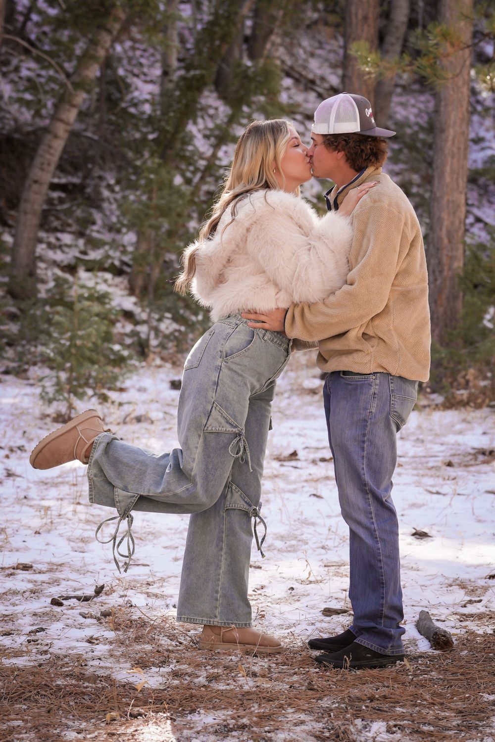 Couple sharing a romantic kiss in a snowy forest, woman in fur jacket and cargo pants, man in tan corduroy shirt and cap.