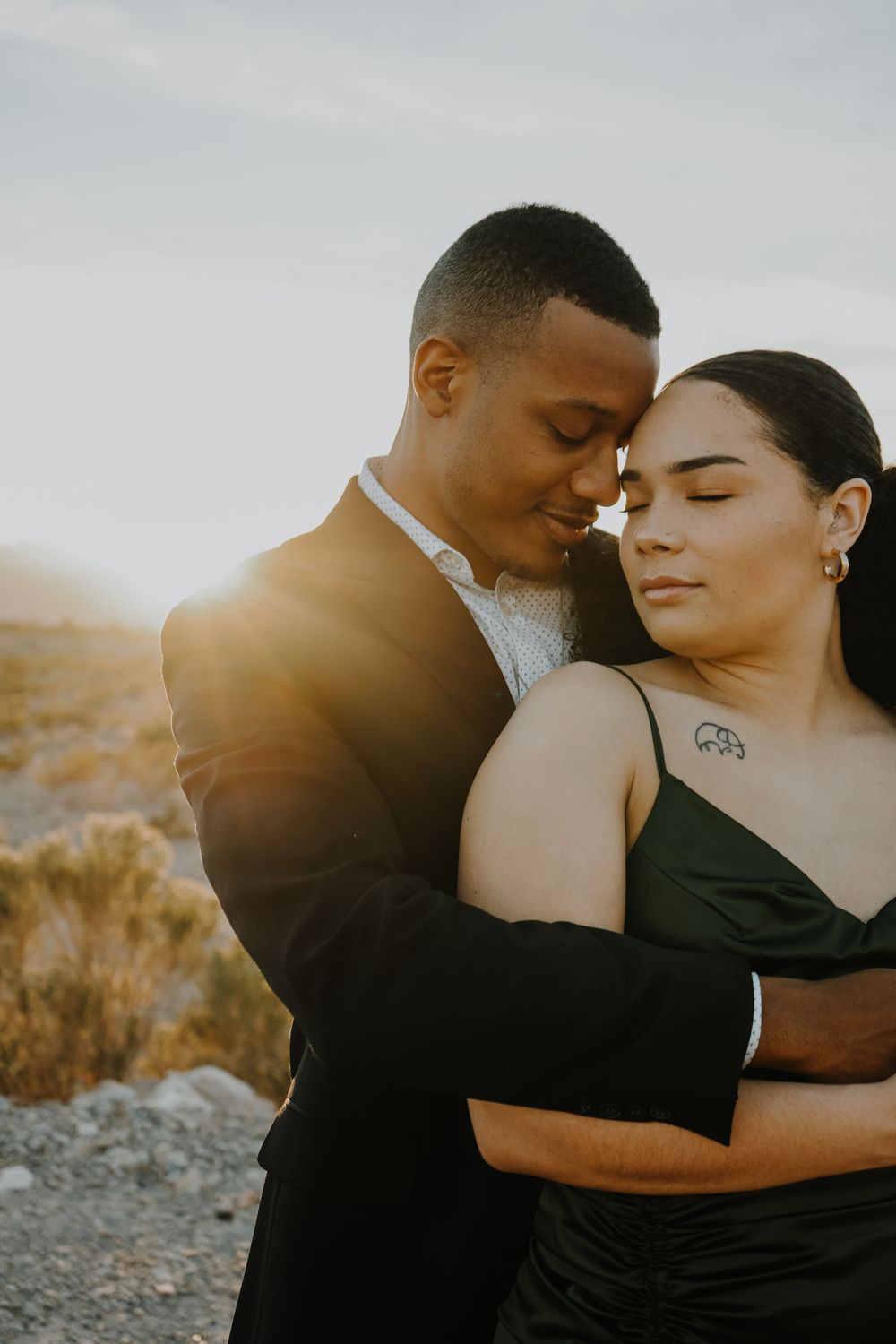 Couple embracing at golden hour in desert, woman in black dress with tattoo, man in dark suit, romantic engagement photo.