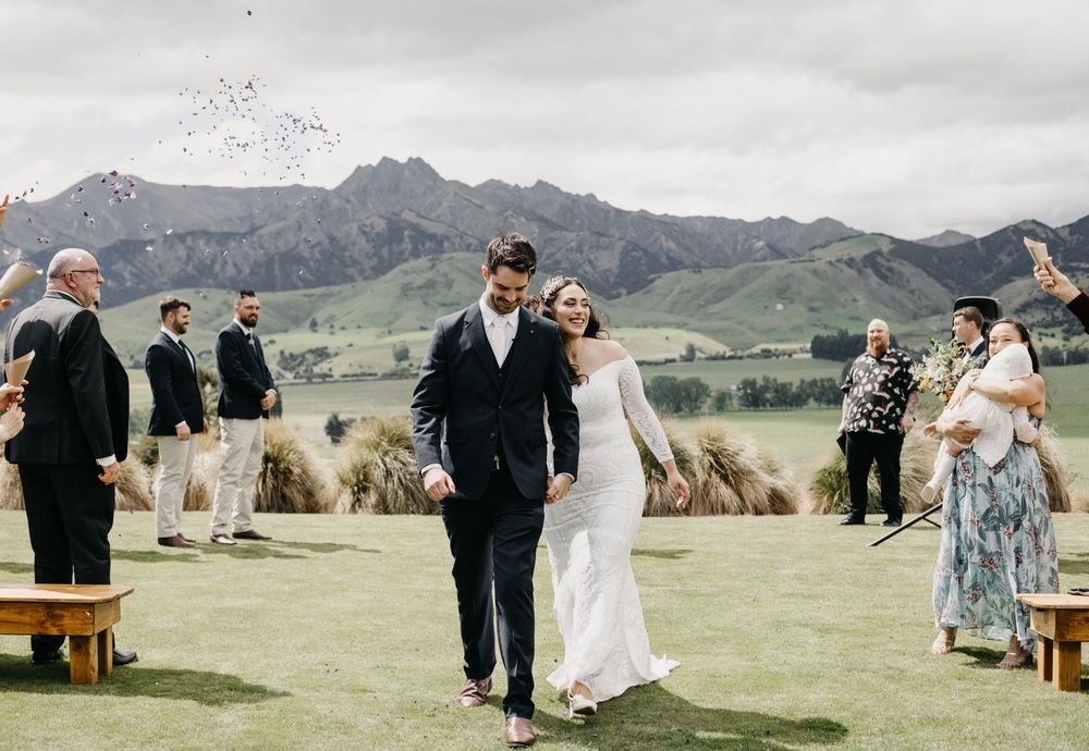 Joyful newlyweds walking through confetti shower with mountain views and wedding guests cheering.