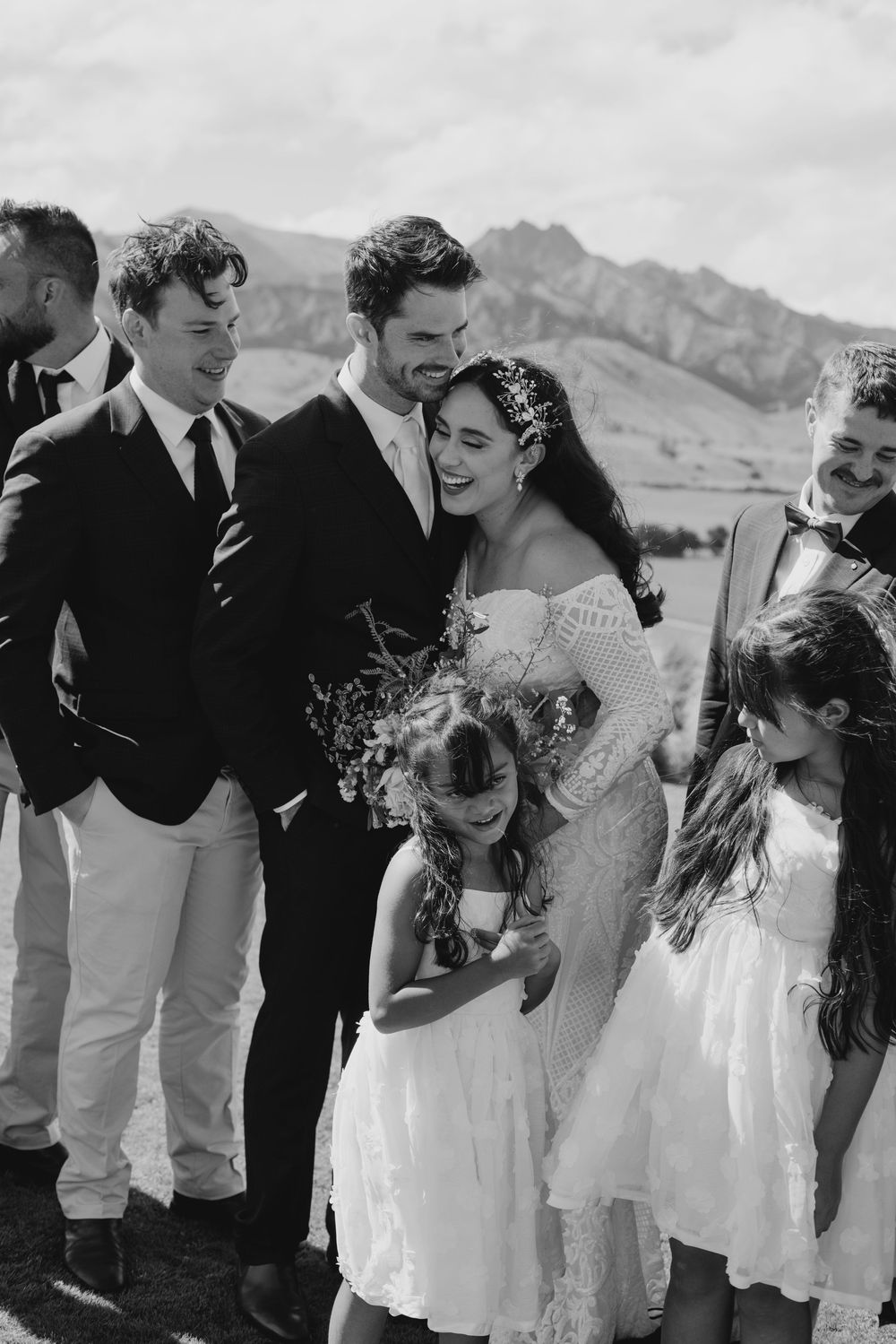 Black and white wedding photo of couple celebrating with guests and flower girls outdoors.