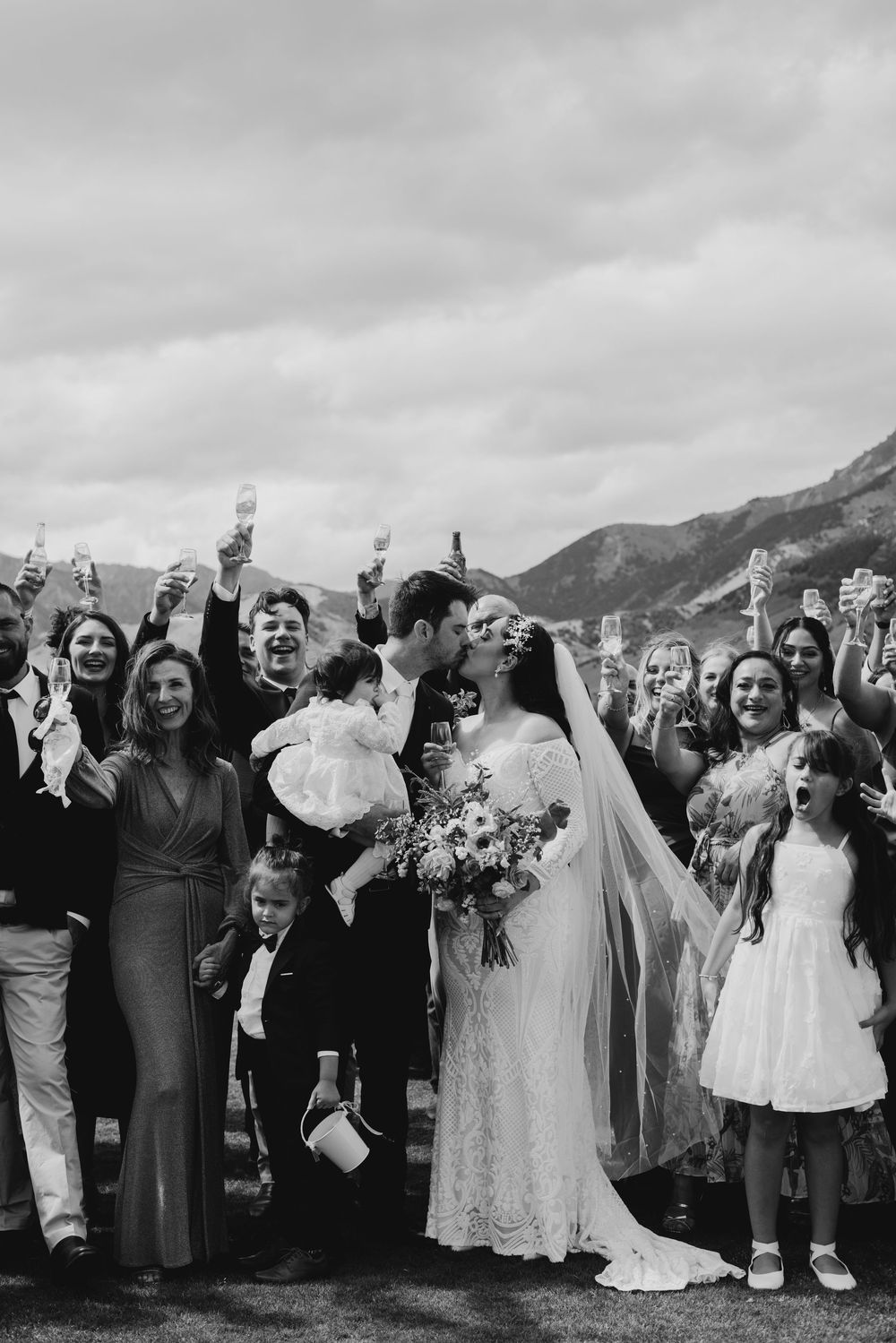 Joyful wedding guests raising glasses to celebrate bride and groom kissing in black and white.