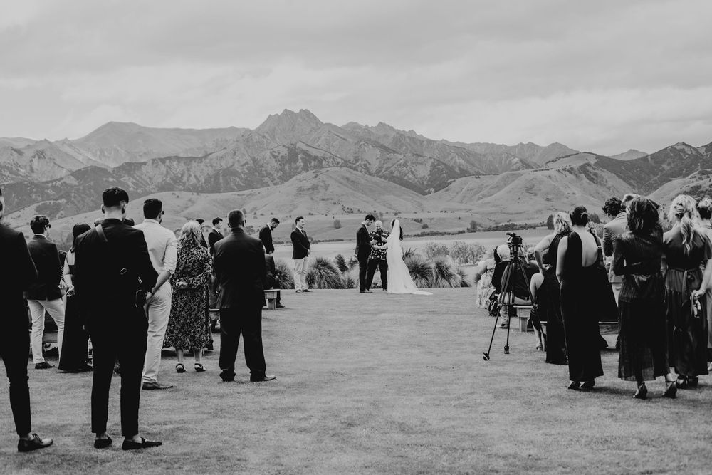 Black and white wide-angle wedding ceremony outdoors with mountain landscape and guests standing.