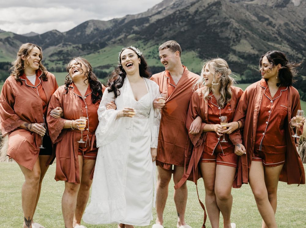 Bride in white robe laughing with bridesmaids in rust satin sets against mountain backdrop.