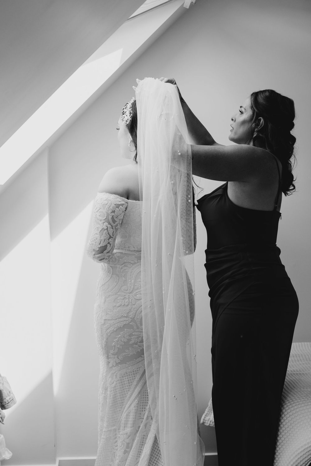 Bridesmaid carefully arranging cathedral veil on bride in lace wedding gown in black and white photo.