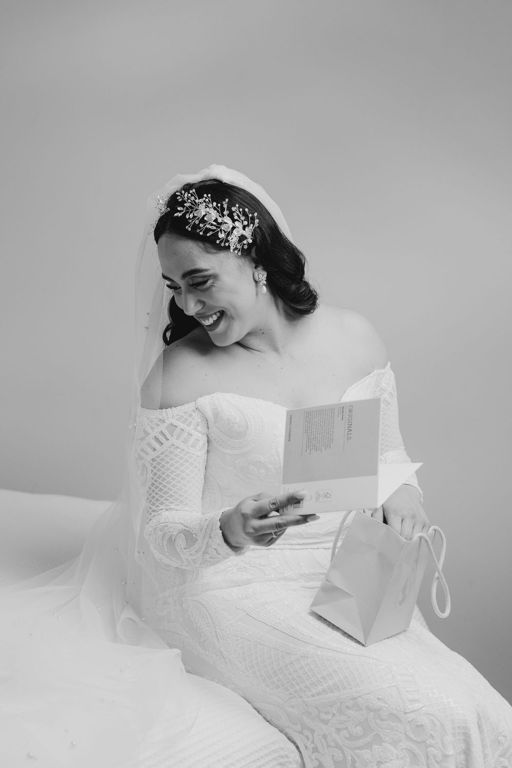 Radiant bride in lace off-shoulder gown reading a card while wearing floral headpiece and veil.