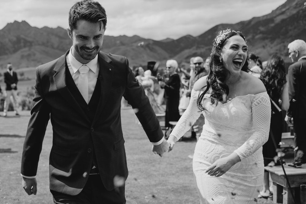 Laughing newlyweds holding hands walking back down aisle after ceremony with mountains in background.