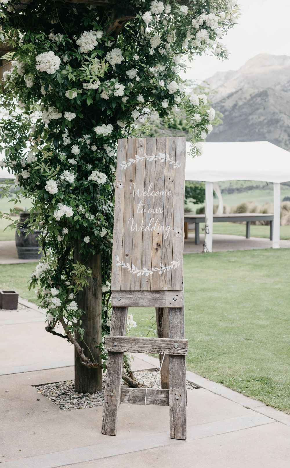 Rustic wooden welcome wedding sign on ladder easel surrounded by white floral greenery.