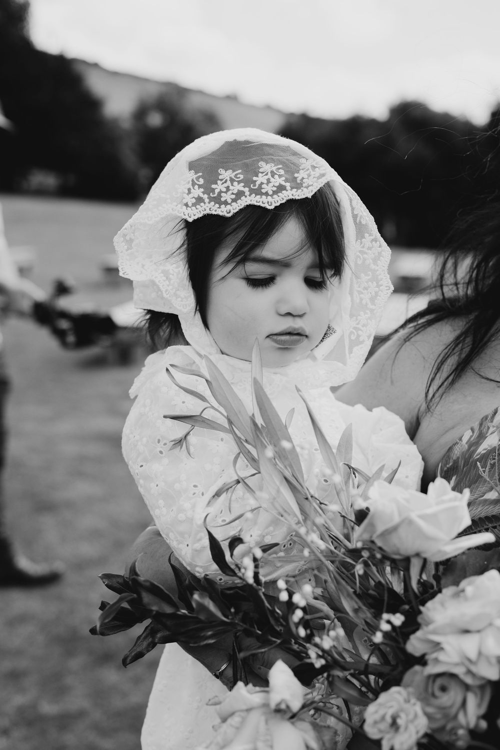 Adorable flower girl in lace veil gazing at bridal bouquet in black and white wedding portrait.