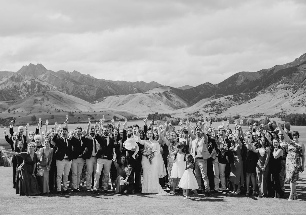 Large wedding party celebrating with raised arms in black and white photo with mountain landscape.