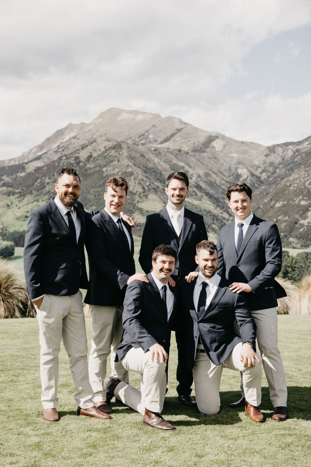 Six groomsmen in navy suits posing together on grass with dramatic mountain range behind them.
