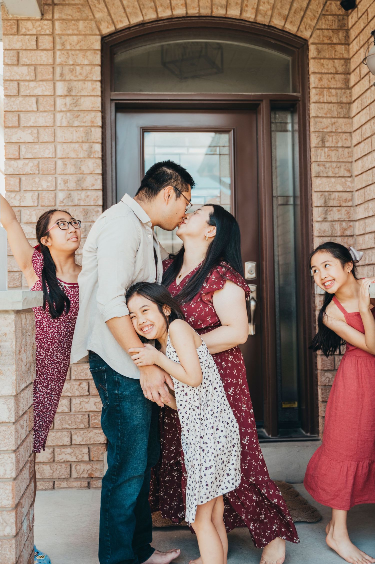 Mom and Dad share a kiss on their front porch while their three children are surrounding them, playing and smiling.