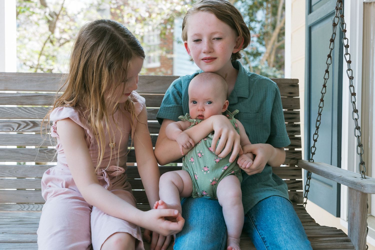 Two children sit on a porch swing holding a baby in a green romper on a sunny day.